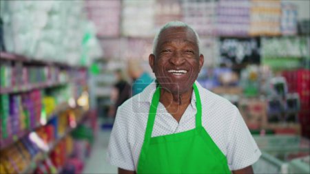 Téléchargez les photos : Un heureux employé noir plus âgé du magasin local souriant à la caméra. Afro-Américain cadre supérieur de l'épicerie portant tablier avec une émotion joyeuse, concept d'emploi salarié - en image libre de droit