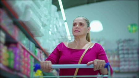 Téléchargez les photos : Femme d'âge moyen naviguant dans l'allée du supermarché, naviguant sur les produits d'étagère, affichant les habitudes d'achat des consommateurs - en image libre de droit