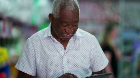 Foto de Un alegre anciano negro brasileño hombre usando dispositivo de tableta. Persona afroamericana mayor comprometida con la tecnología moderna, mirando la pantalla sonriendo - Imagen libre de derechos