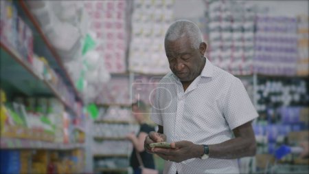 Téléchargez les photos : Un Brésilien aîné noir vérifiant téléphone debout à l'intérieur de l'épicerie allée notes de navigation sur smartphone appareil - en image libre de droit
