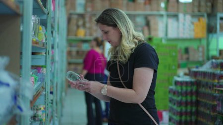 Téléchargez les photos : Consumer in Dilemma, Picking and Comparing Products at Store Shelf. Femme inspectant tasse en verre à l'entreprise locale - en image libre de droit