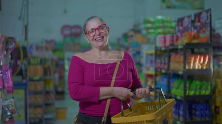 Téléchargez les photos : Heureux client féminin tenant panier debout à l'intérieur du supermarché. Femme d'âge moyen souriant à l'épicerie représentant habitudes de vie consumérisme - en image libre de droit