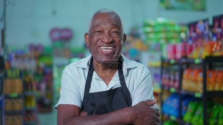 Photo for Portrait of a joyful African American senior employee of supermarket wearing apron and smiling at camera inside grocery store aisle and arms crossed - Royalty Free Image