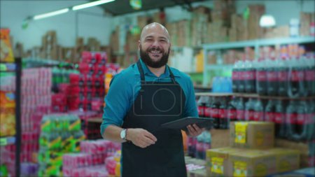 Foto de Retrato de un alegre empleado brasileño de supermercado parado en el pasillo con delantal y dispositivo de tableta en la mano - Imagen libre de derechos