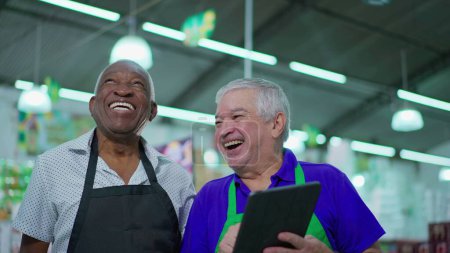 Téléchargez les photos : Confrères aînés heureux de supermarché riant et souriant, interaction joyeuse authentique franche entre les hommes âgés du personnel diversifié tout en tenant tablette dispositif - en image libre de droit