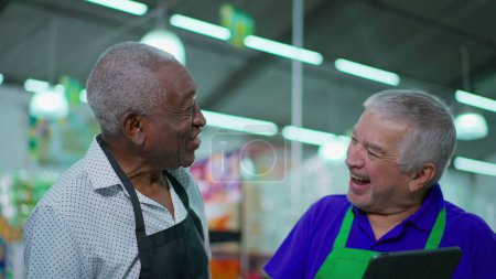 Foto de Felices colegas de alto nivel del supermercado riendo y sonriendo, sincera interacción alegre auténtica entre los hombres de más edad personal diverso mientras sostiene el dispositivo de la tableta - Imagen libre de derechos