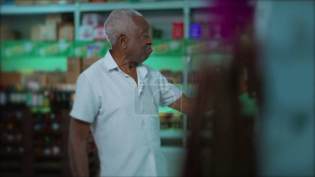 Photo for Candid elderly African American man selecting beverage from shelf at grocery store. One senior black male shopping for alcoholic drink at supermarket - Royalty Free Image