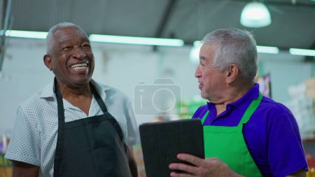 Téléchargez les photos : Confrères aînés heureux de supermarché riant et souriant, interaction joyeuse authentique franche entre les hommes âgés du personnel diversifié tout en tenant tablette dispositif - en image libre de droit