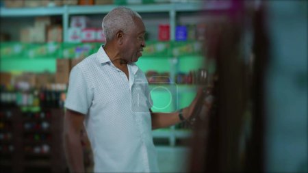 Photo for Candid elderly African American man selecting beverage from shelf at grocery store. One senior black male shopping for alcoholic drink at supermarket - Royalty Free Image