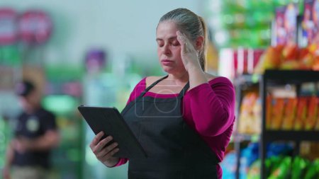 Foto de Empleada mujer ansiosa usando tableta de pie en el supermercado lamentando error. Personal de mediana edad poniendo la mano en la frente se siente frustrado mientras se utiliza la tecnología moderna - Imagen libre de derechos