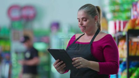 Foto de Empleada mujer ansiosa usando tableta de pie en el supermercado lamentando error. Personal de mediana edad poniendo la mano en la frente se siente frustrado mientras se utiliza la tecnología moderna - Imagen libre de derechos
