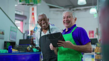 Photo for Joyful diverse Brazilian senior staff workers of supermarket chain smiling at camera with table and uniforms. African American older employee and a caucasian person laughing together - Royalty Free Image