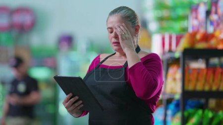 Photo for Anxious female employee using tablet standing in supermarket regretting mistake. Middle-aged staff putting hand in forehead feeling frustrated while using modern technology - Royalty Free Image