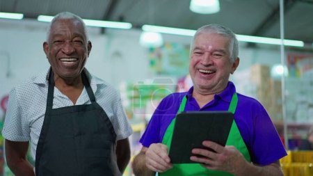 Téléchargez les photos : Candid Authentic Joy Between Older Supermarket Colleagues doing high-five, Souriant Divers hommes âgés avec tablette dispositif, les travailleurs célébrant le succès - en image libre de droit
