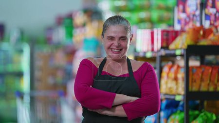 Foto de Mujer alegre trabajadora de la tienda de comestibles sonriendo a la cámara con los brazos cruzados, usando delantal - Imagen libre de derechos