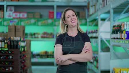 Téléchargez les photos : Joyeuse employée du supermarché debout à l'allée avec les bras croisés, expression joyeuse d'une employée portant un tablier à l'épicerie - en image libre de droit