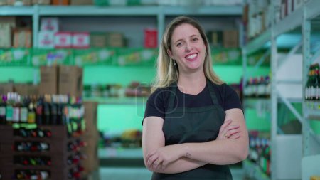 Foto de Feliz empleada del supermercado parada en el pasillo con los brazos cruzados, expresión alegre de una mujer que usa delantal en la tienda de comestibles - Imagen libre de derechos