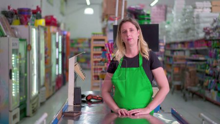 Téléchargez les photos : Employée de Supermarché debout à la caisse en regardant la caméra avec expression neutre portant tablier vert. Portrait du personnel de l'épicerie - en image libre de droit
