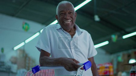 Téléchargez les photos : Un heureux acheteur brésilien âgé souriant à la caméra avec panier à l'intérieur du hangar du supermarché. Portrait d'un consommateur afro-américain plus âgé à l'épicerie - en image libre de droit
