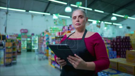 Téléchargez les photos : Employée sérieuse d'âge moyen d'un supermarché debout à l'intérieur d'une entreprise tenant un appareil tablette, regardant la caméra avec une expression neutre - en image libre de droit