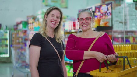 Photo for Two joyful female shoppers smiling at camera inside Supermarket store holding basket in hand. Portrait faces of smiling middle-aged women doing grocery shopping - Royalty Free Image