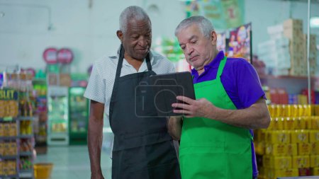 Foto de Dos altos empleados diversos que trabajan en el supermercado mirando el dispositivo de la tableta, escena del trabajo en equipo del gerente más viejo orientando al empleado en la tienda de comestibles, concepto de ocupación laboral - Imagen libre de derechos