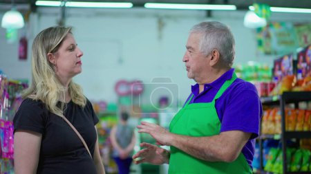 Foto de Empleada senior de la tienda de comestibles hablando con una clienta dentro del supermercado. Mayor gerente usando delantal conversando con la mujer - Imagen libre de derechos