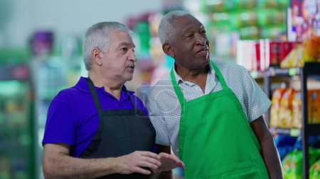 Photo for Senior diverse employees of grocery store discussing management of business at supermarket aisle wearing uniforms - Royalty Free Image
