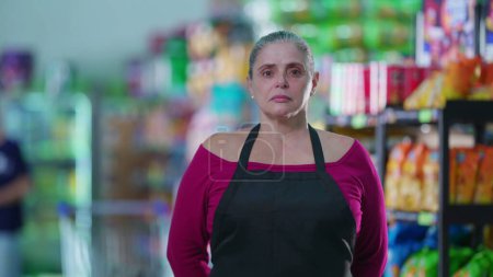Téléchargez les photos : Travailleuse d'âge moyen préoccupée par le portrait de supermarché debout dans l'allée avec une expression inquiète, portant un tablier - en image libre de droit