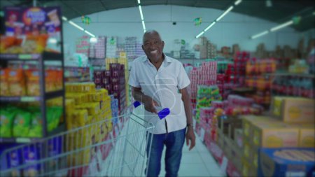 Téléchargez les photos : Souriant afro-américain âgé Shopper avec chariot dans l'allée du supermarché - en image libre de droit