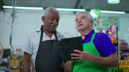 Téléchargez les photos : Deux cadres supérieurs heureux et diversifiés de la glacerie vérifient les opérations d'inventaire avec un appareil tablette, les employés plus âgés du supermarché rient et sourient au travail - en image libre de droit
