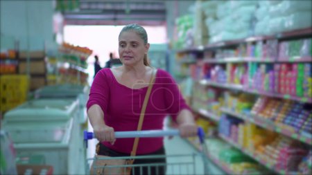 Téléchargez les photos : Femme d'âge moyen faisant du shopping dans un supermarché marchant à travers les produits de navigation allée sur étagère, Affichage du consumérisme - en image libre de droit