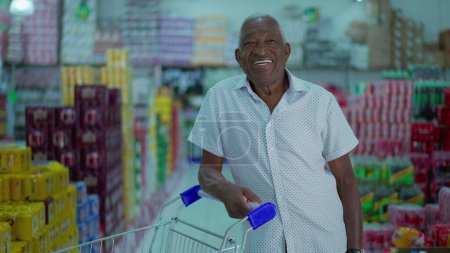 Foto de Comprador afroamericano sonriendo a la cámara de pie dentro del supermercado con el carrito de compras. Un consumidor negro brasileño en la tienda de comestibles - Imagen libre de derechos