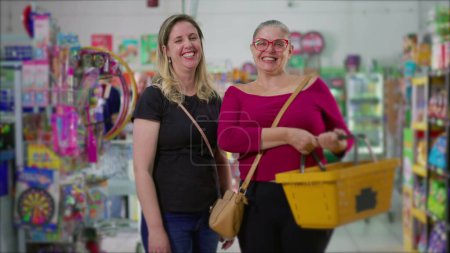 Foto de Dos clientes felices hembras de pie dentro de la tienda de comestibles sosteniendo la cesta en la mano. Retrato de las mujeres compradoras en el supermercado sonriendo - Imagen libre de derechos