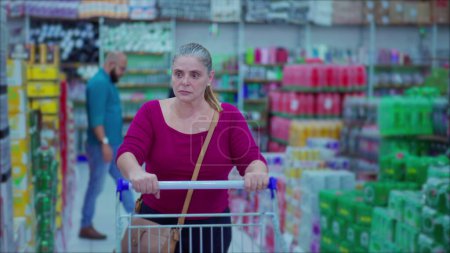 Téléchargez les photos : Femme shopper poussant panier marchant dans les produits de navigation allée supermarché représentant les habitudes de vie consumérisme. Consommation féminine d'âge moyen - en image libre de droit