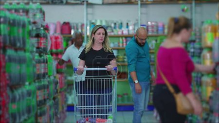 Téléchargez les photos : Femme poussant chariot d'achat à l'épicerie allée avec des acheteurs en arrière-plan représentant les habitudes de consommation à l'épicerie - en image libre de droit