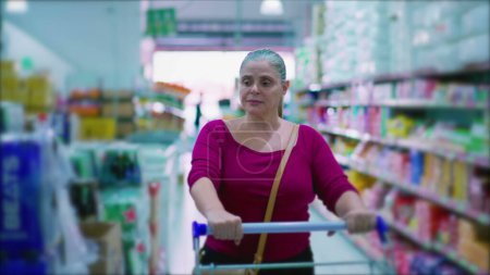 Téléchargez les photos : Femme consommateur marchant dans un supermarché à la recherche de produits à acheter, femme acheteur poussant panier d'achat à l'épicerie - en image libre de droit