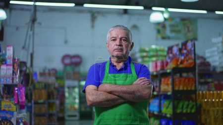 Téléchargez les photos : Gestionnaire de supermarché sérieux avec bras croisés, expression préoccupée de l'ancien propriétaire de petite entreprise à l'intérieur du magasin - en image libre de droit