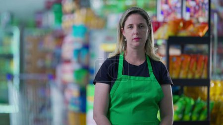 Téléchargez les photos : Employée sérieuse du supermarché portant un tablier vert, personnel d'âge moyen de l'épicerie avec une expression craintive sévère pendant les périodes difficiles, profession - en image libre de droit