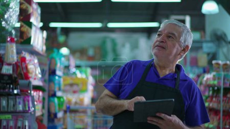 Foto de Gerente senior de la tienda de comestibles revisando el inventario de productos en el estante con el dispositivo de la tableta, empleado masculino caucásico inspeccionando artículos en el pasillo del supermercado - Imagen libre de derechos