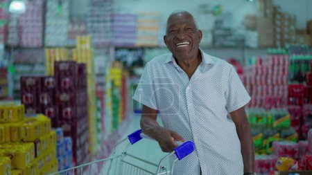 Foto de Comprador afroamericano sonriendo a la cámara de pie dentro del supermercado con el carrito de compras. Un consumidor negro brasileño en la tienda de comestibles - Imagen libre de derechos