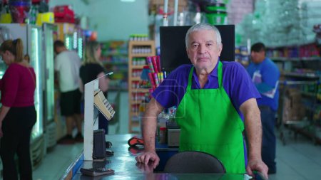 Photo for Senior employee of Supermarket standing by cashier checkout with neutral serious expression. older staff of grocery store with business and customers in background - Royalty Free Image