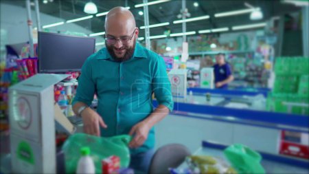 Téléchargez les photos : Un employé brésilien heureux de supermarché à la caisse des produits de numérisation et de mettre dans des sacs en plastique tout en souriant à la caméra. Marché de l'épicerie en Amérique du Sud - en image libre de droit
