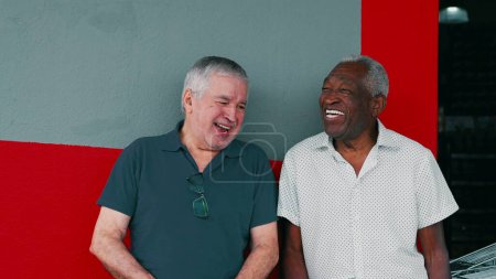 Foto de Joyful Interaction of Two Diverse Male Senior Friends, Smiling and Laughing Together, Leaning on Sidewalk Wall (en inglés). Auténticos ancianos felices de la vida real - Imagen libre de derechos