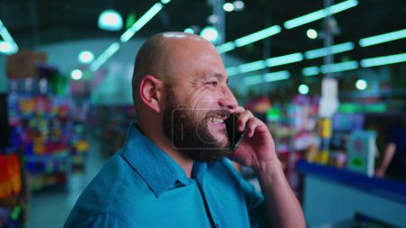 Téléchargez les photos : Homme barbu heureux ayant conversation téléphonique dans un magasin sud-américain hangar, individu occasionnel avec Smartphone moderne - en image libre de droit