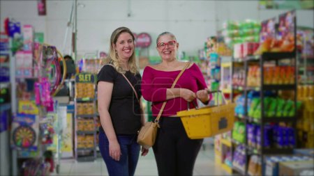 Téléchargez les photos : Deux clientes heureuses debout à l'intérieur de l'épicerie tenant le panier à la main. Portrait de femmes acheteurs au supermarché souriant - en image libre de droit
