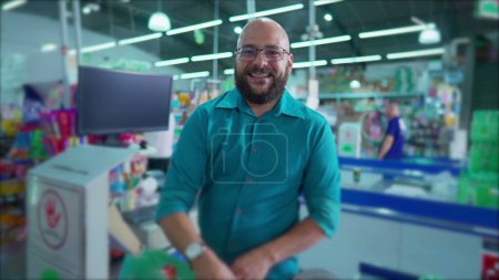 Foto de Un empleado brasileño feliz del supermercado en la compra de productos de escaneo y poner en bolsas de plástico mientras sonríe a la cámara. Escena del mercado de comestibles en América del Sur - Imagen libre de derechos