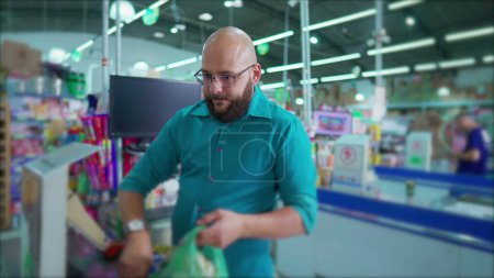 Téléchargez les photos : Joyeux employé du supermarché brésilien à la caisse Scanning and Bagging Products, souriant à la caméra sur la scène du marché sud-américain de l'épicerie - en image libre de droit