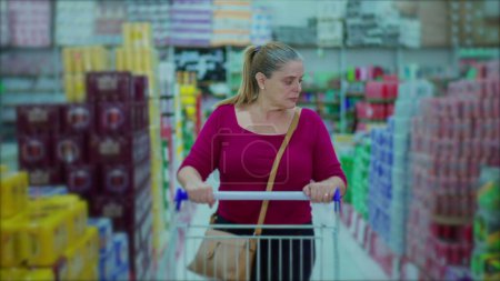 Téléchargez les photos : Femme marchant dans les produits de navigation allée supermarché sur étagère. Acheteur d'âge moyen poussant le panier - en image libre de droit
