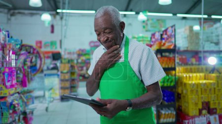Foto de Empleado afroamericano mayor confundido con tableta dentro de la tienda de comestibles, miembro mayor del personal que intenta navegar por la tecnología - Imagen libre de derechos